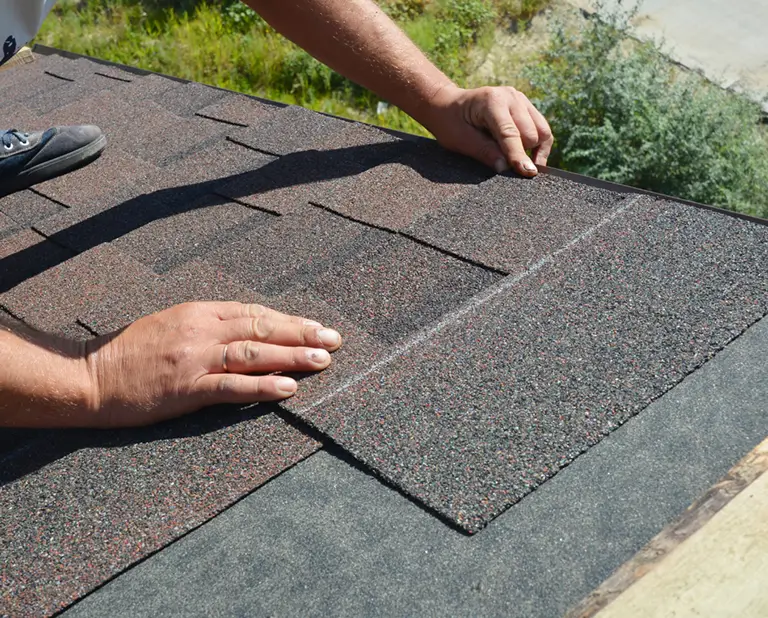 Hands of a person installing asphalt shingles on a roof. The shingles are dark gray with a textured surface, set against a sunny outdoor backdrop.