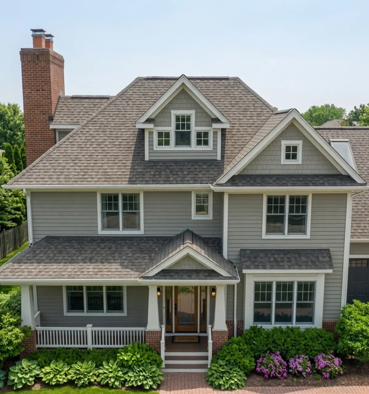 Two-story gray house with white trim, featuring a steep roof, front porch, and brick chimney. Lush green bushes and purple flowers line the walkway.