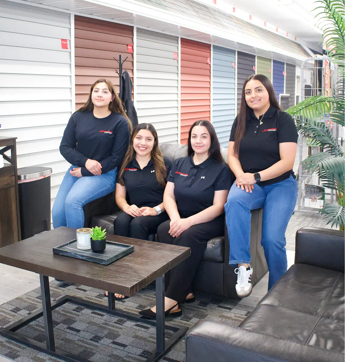 Four women sit and stand on a leather couch in a showroom, wearing branded uniforms. Behind them are colorful wall panels. The setting is modern and professional.
