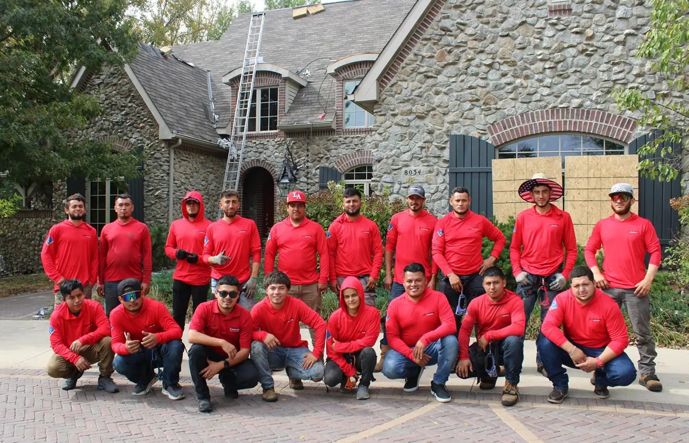 A group of 17 people wearing matching red shirts poses in front of a stone house. A ladder leans against the roof. The mood is team-oriented and lively.