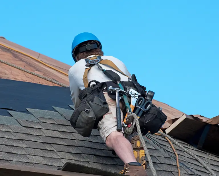 Worker on a roof, wearing a blue helmet and safety harness, adjusts shingles against a clear blue sky. Tool belt and climbing gear are visible.